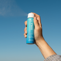 Hand holding a COOLA sunscreen travel size spray bottle SPF50 Fragrance Free against a clear blue sky