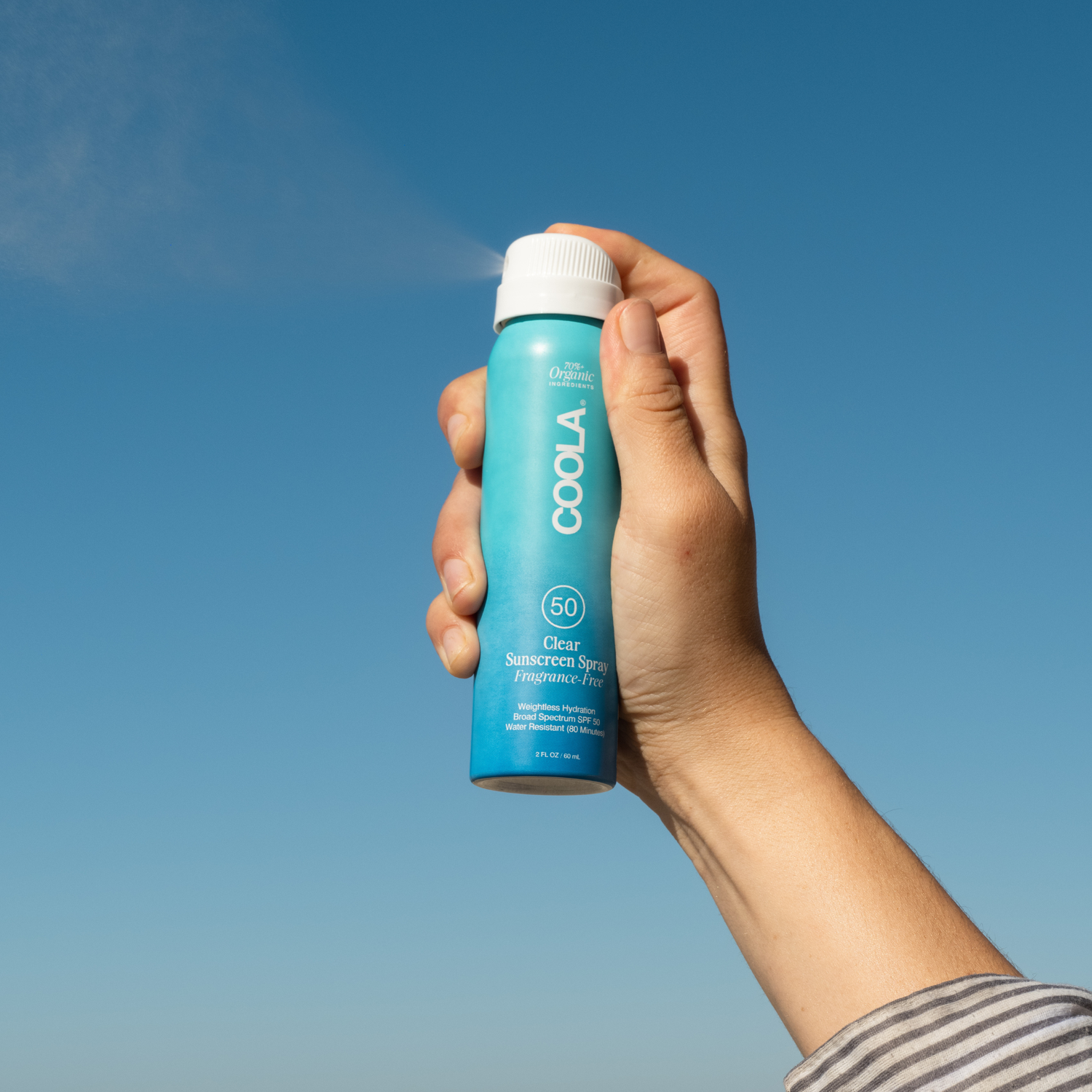 Hand holding a COOLA sunscreen travel size spray bottle SPF50 Fragrance Free against a clear blue sky