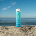 Coola sunscreen travel size spray bottle SPF50 Fragrance Free  on a sandy beach with ocean and blue sky in the background