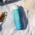 COOLA sunscreen travel size spray bottle SPF50 Fragrance Free on a textured surface with sunglasses and a book.