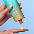 A hand holds a tube of COOLA Hydrating Sunscreen Lotion SPF30 in Tropical Coconut, with the orange cap unscrewed and a dollop of white lotion shown on another hand against a light blue background.