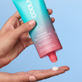 A hand holds a teal-to-pink gradient tube of COOLA Hydrating Sunscreen Lotion (Guava Mango) SPF 50, with white sunscreen cream being squeezed onto another hand against a light blue background.