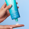 A hand holds a turquoise tube of COOLA Hydrating Sunscreen Lotion SPF50 Fragrance Free, with white sunscreen cream squeezed onto the other hand against a light blue background.