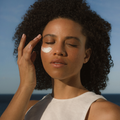 Woman applying Glory Cream to her face with a clear blue sky and ocean in the background