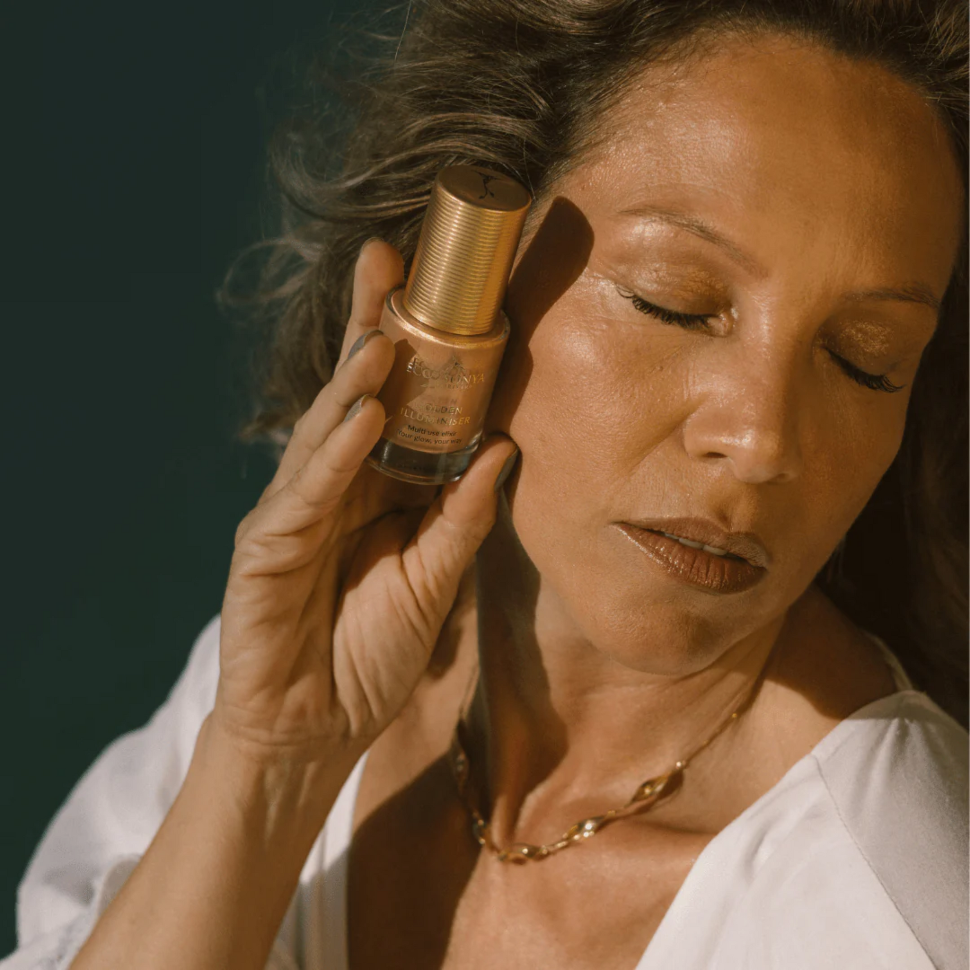 Woman holding a Eco by Sonya Golden Illuminiser bottle close to her ear against a dark background
