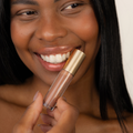 A close-up of a smiling woman holding a clear tube of Eco by Sonya Glory Gloss in Maple 08 color with a gold cap, showing the nude pink shade against a light neutral background.