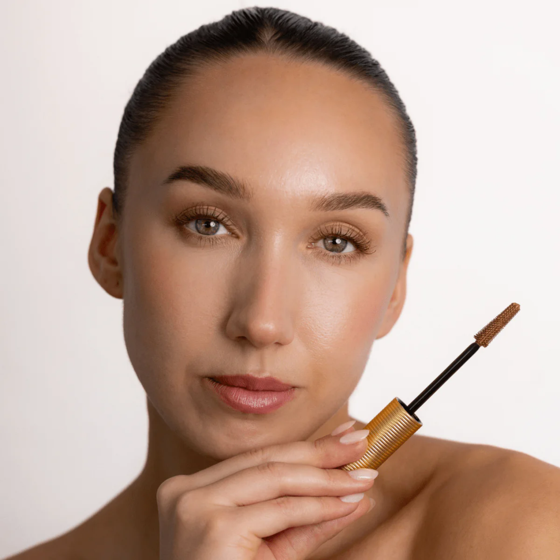 A gold ribbed Eco by Sonya Brown Mascara tube shown open with the wand removed, revealing a black applicator wand with a brown bristle tip, on a plain white background.