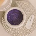 Purple super acai exfoliator  in a white container on a textured beige surface with a spoon labeled 'Mix & Scoop'.