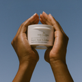 Hands holding a jar of Eco by Sonya Super Acai Exfoliant against a clear blue sky.