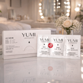YUMI lashes and brow 3.0 products on a reflective surface with a blurred bedroom background