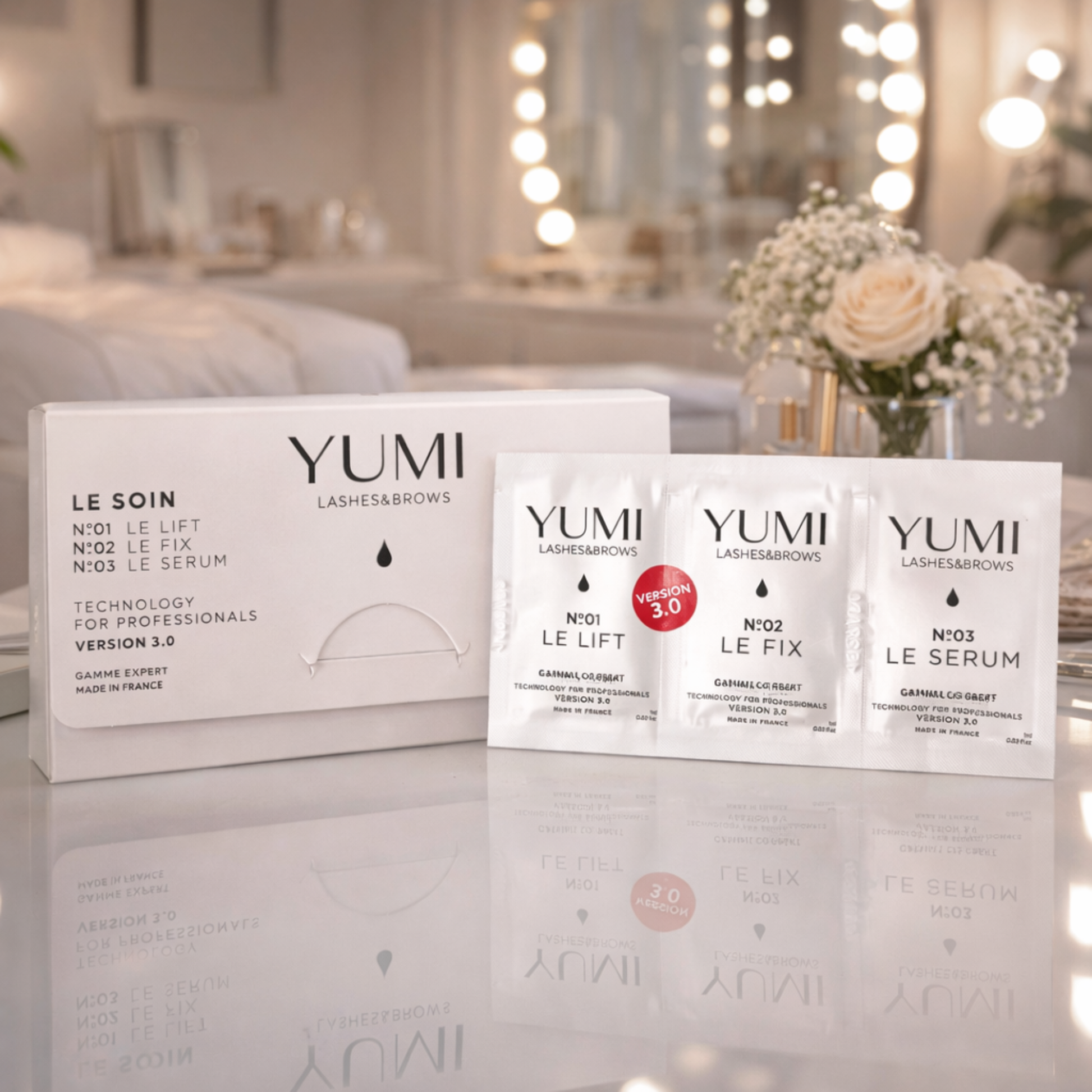 YUMI lashes and brow 3.0 products on a reflective surface with a blurred bedroom background