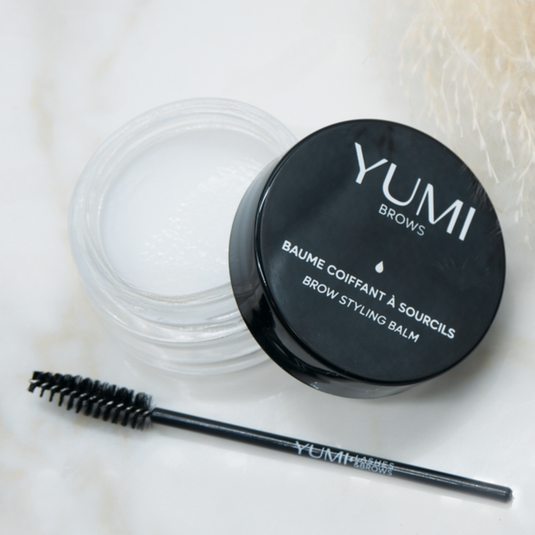 Yumi Brows Brow Styling Balm in a small jar, showcasing the product label and the dark brown color of the balm.